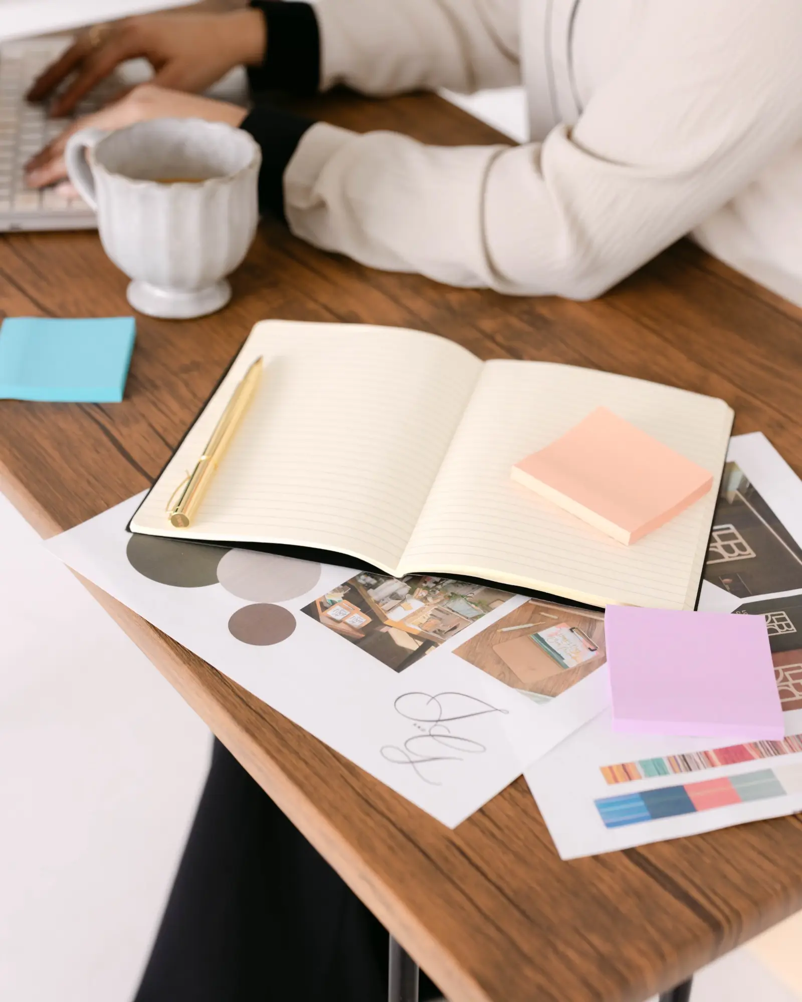 An open notebook and gold pen on brand mood board prints, beside a cup of tea and a laptop